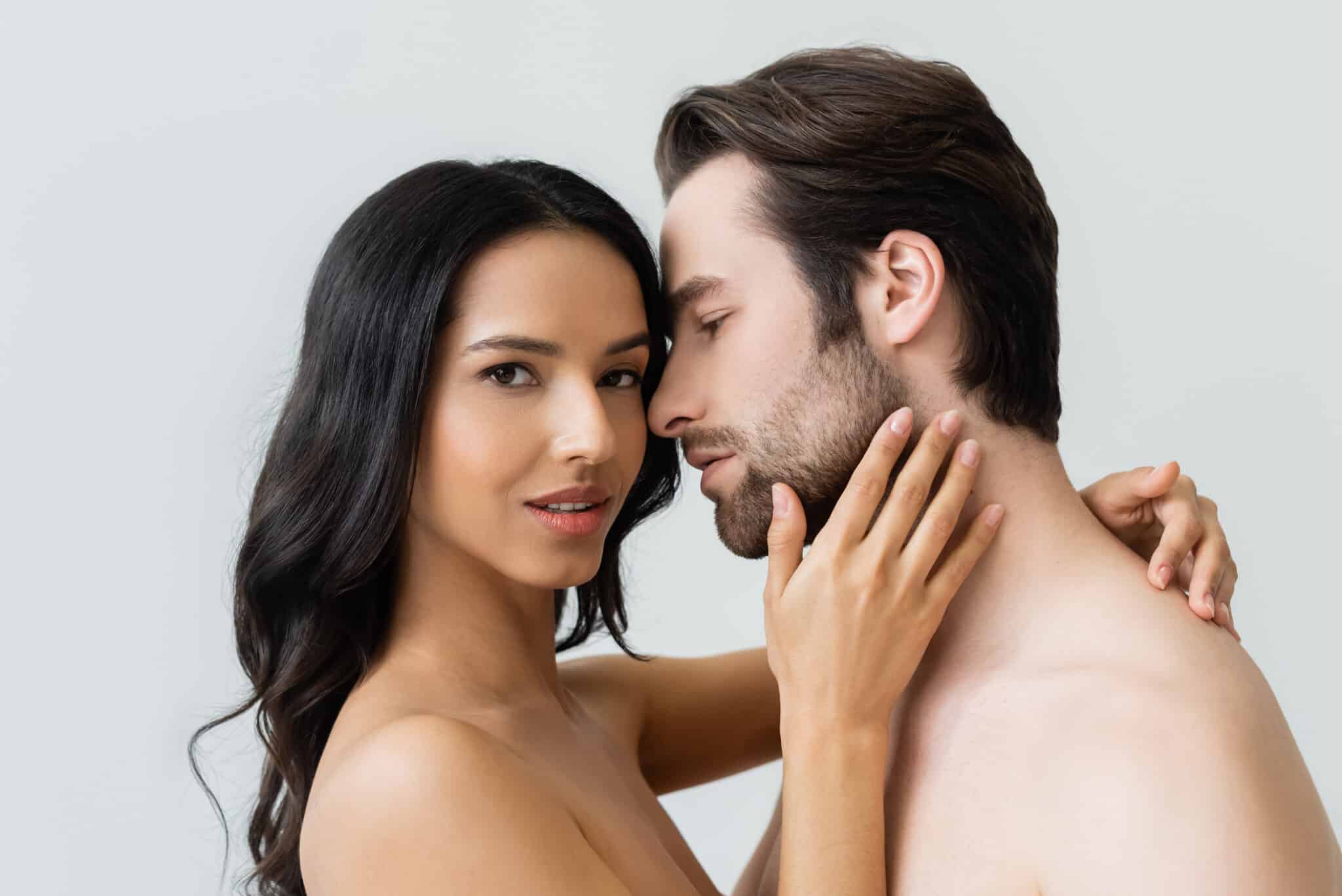 young and sensual woman looking at camera while hugging bearded man isolated on grey